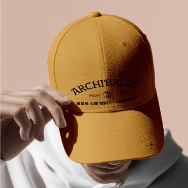 Person adjusting a mustard-coloured Archibald’s branded cap in soft natural light