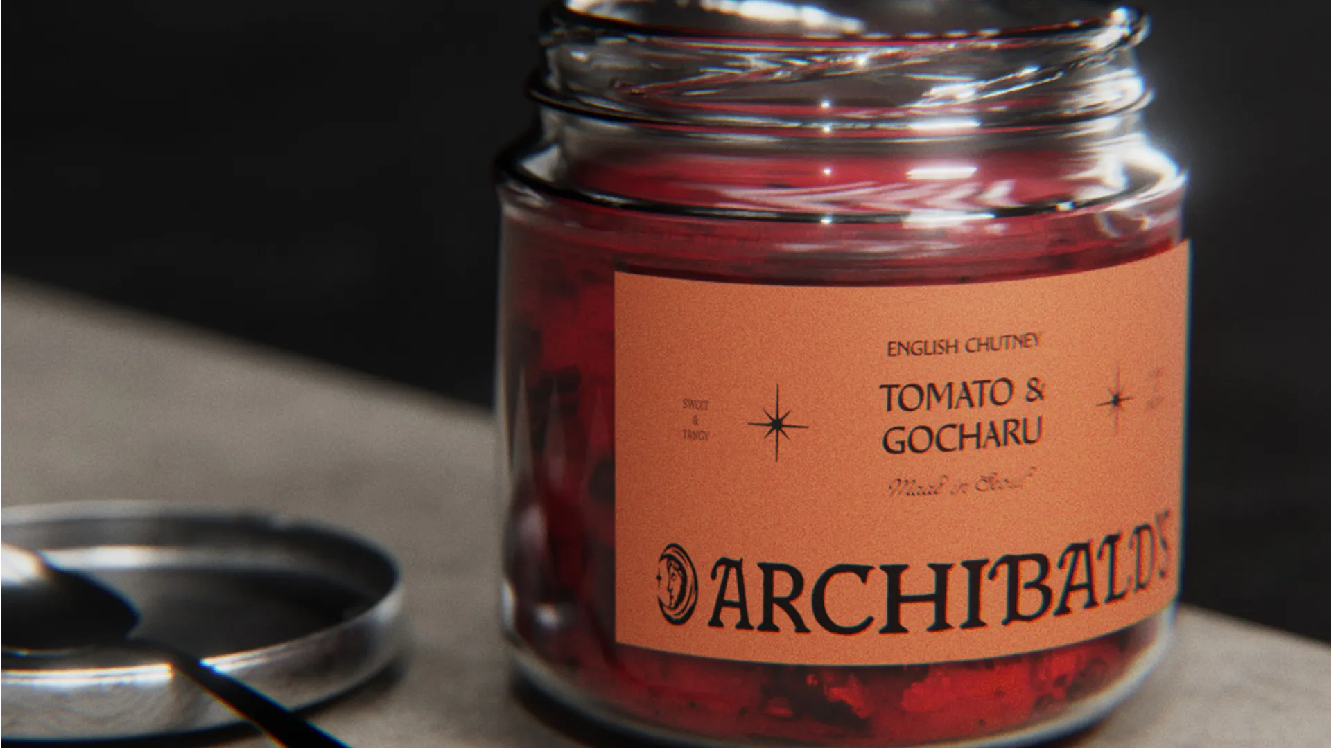 Glass jar of Archibald’s Tomato & Gocharu chutney beside its lid and a small spoon