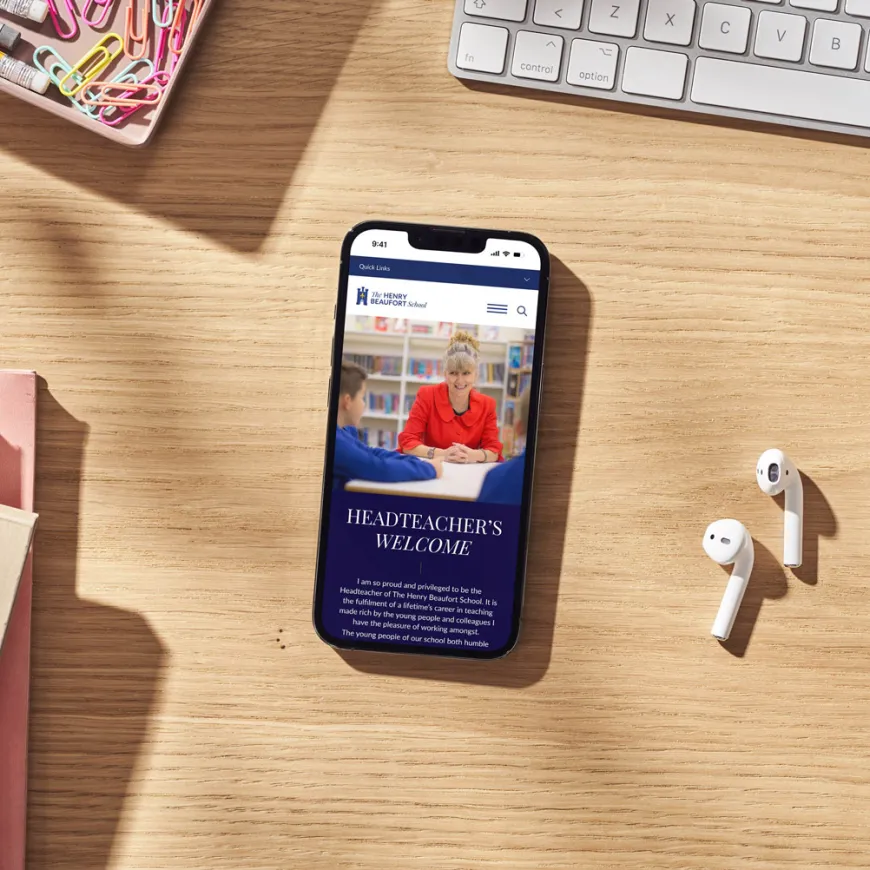 Smartphone showing the school's website on a wooden desk beside stationery, earphones and a coffee cup